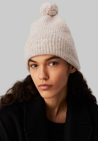 Berretto in maglia beige con una texture a coste e un pom-pom in cima. Presenta un piccolo logo ricamato sul davanti. Materiale in morbida lana.