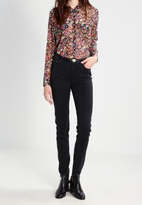 Kvinna i en blommig skjorta med långa ärmar, svarta tajta jeans och svarta ankelstövlar, stående med en hand i fickan.