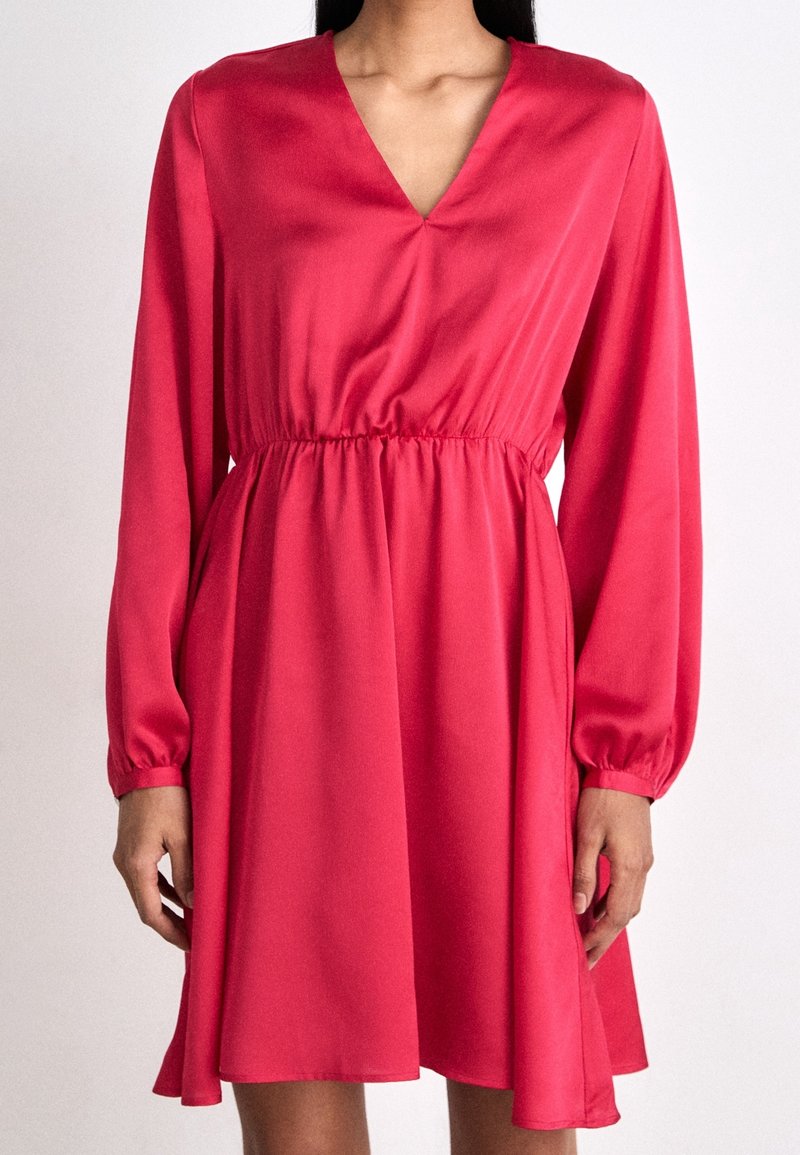Femme portant une robe rouge à manches longues, longueur genou, col en V et taille froncée.