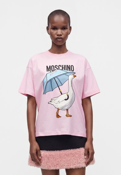 Γυναίκα που φορά ένα ανοιχτό ροζ T-shirt Moschino με ένα γραφικό λευκής χήνας που κρατά μια μπλε ομπρέλα.