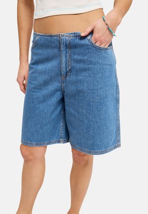 Personne portant un short en jean bleu taille mi-haute, ample, avec des poches avant et un haut court blanc, main placée dans la poche droite.