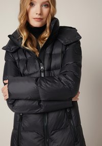 Kobieta z długimi blond włosami, ubrana w czarn�ą ocieplaną zimową kurtkę z wysokim kołnierzem, z założonymi na siebie rękami i zamkami błyskawicznymi w przednich kieszeniach.