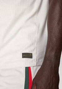 Maglia sportiva bianca con strisce verticali testurizzate, una patch del logo sull'orlo e accenti laterali colorati in verde e rosso.