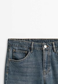 Gros plan sur la ceinture d'un jean en denim bleu avec bouton avant, passants pour ceinture et détails de la poche avant sur fond blanc.