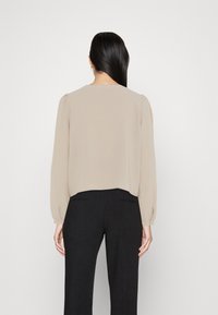 Femme aux cheveux longs et foncés portant un chemisier beige à manches longues et un pantalon noir, debout face à un fond blanc uni.