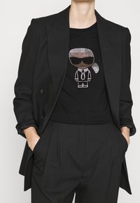 Person som bär en svart kavaj och byxor med händerna i fickorna och en svart t-shirt med en strassdekorerad tecknad figur som bär solglasögon och slips.