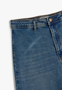 Jeans de denim en azul medio con una textura desgastada, que cuentan con un diseño tradicional de cinco bolsillos, cierre de botón y trabillas para el cinturón.