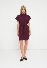 Βουργουνδί φόρεμα με κοντά μανίκια, στενεμένο στη μέση με ζωνάκι, φτιαγμένο από μαλακό ύφασμα. Μαύρα μυτερά παπούτσια ολοκληρώνουν την εμφάνιση.