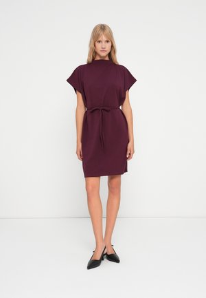 Burgundin mekko, jossa on lyhyet hihansuut, vyötäröllä solmittuna, valmistettu pehmeästä kankaasta. Mustat teräväkärkiset kengät täydentävät asua.