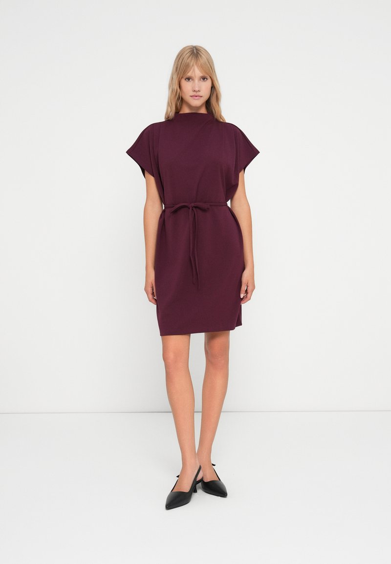 Βουργουνδί φόρεμα με κοντά μανίκια, στενεμένο στη μέση με ζωνάκι, φτιαγμένο από μαλακό ύφασμα. Μαύρα μυτερά παπούτσια ολοκληρώνουν την εμφάνιση.