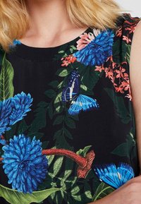 Femme portant un haut sans manches noir avec des fleurs bleues, des feuilles vertes et des papillons bleus imprimés sur le tissu.