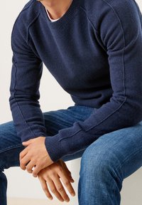 Homme portant un pull bleu marine et un jean bleu, assis les mains croisées et reposant sur ses genoux contre un fond uni.
