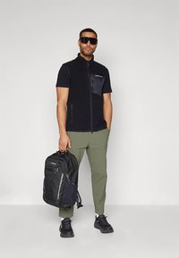 Črna telovnik z zadrgo z teksturiranim žepom, olivno zelene hlače, črn nahrbtnik in superge. Eleganten dizajn s funkcionalnimi podrobnostmi.