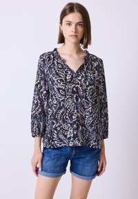 Junge Frau mit glattem braunem Haar, die eine dunkle gemusterte Bluse mit 3/4-Ärmeln und hochgekrempelten blauen Jeansshorts vor einem einfarbigen Hintergrund trägt.