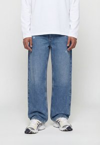 Άνδρας που φοράει μπλε τζιν σε φαρδιά γραμμή, άσπρο μακρυμάνικο πουκάμισο και άσπρα αθλητικά παπούτσια, στέκεται μπροστά σε απλή φόντο.