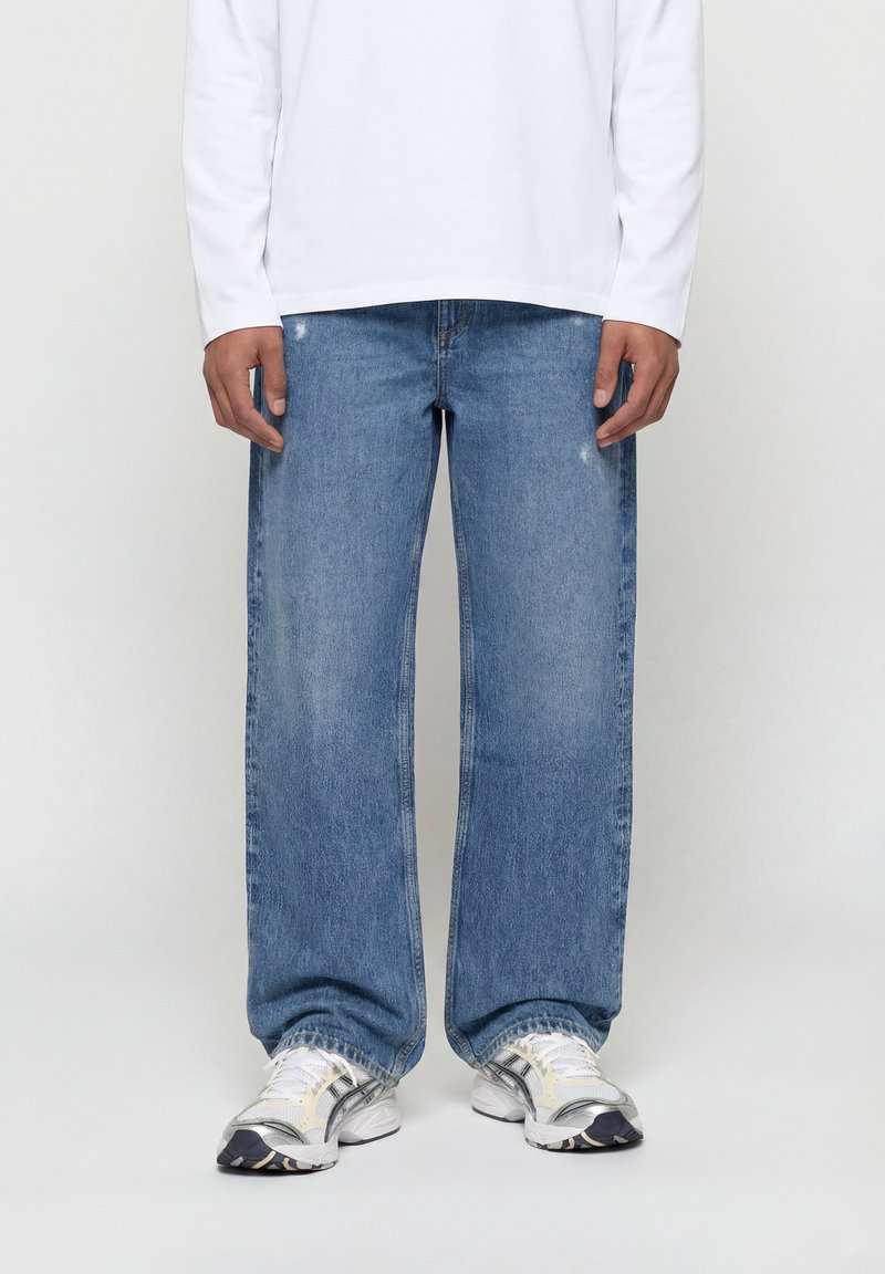 Άνδρας που φοράει μπλε τζιν σε φαρδιά γραμμή, άσπρο μακρυμάνικο πουκάμισο και άσπρα αθλητικά παπούτσια, στέκεται μπροστά σε απλή φόντο.