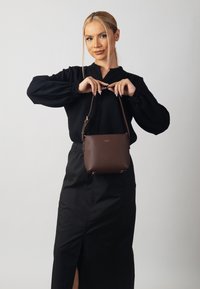 Brown leather handbag with a textured finish, featuring a short strap and gold-tone hardware, held by a person in a black blouse.