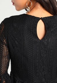 Kvinna i svart spetsklänning med nyckelhålsrygg och knäppning, samt stora guldfärgade hoopsörhängen, visad från axel uppåt i profil.