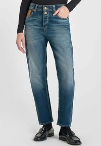 Jeans de denim azul con corte recto, con un lavado desgastado, dos bolsillos frontales, botones de metal y una etiqueta de cuero marrón en la parte trasera.