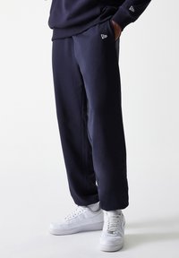 Marineblå sweatpants lavet af blødt stof, med sidelommer og et lille logo på hoften, parret med hvide højtop sneakers.