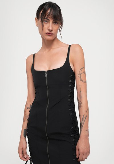 Woman with dark hair and tattoos wearing a black dress with front zipper and side lace-up details against a plain light background.