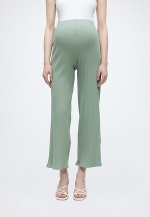 Personne enceinte portant un pantalon de maternité côtelé vert clair, un débardeur blanc et des sandales blanches à brides sur un fond uni.