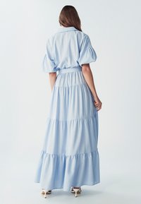 Světle modré maxi šaty s límečkem, vyfouknutými rukávy a vrstvenými volánky, vyrobené z měkké tkaniny. Stylizované s bílými podpatky.