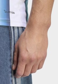 Detailní záběr na uvolněnou ruku vedle stehna, oblečenou v modrých džínách a bílo-modré pruhované tričko climacool.