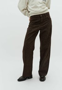 Braune Cordhosen mit einem Zebra-Streifen-Muster, ausgestattet mit einem Knopf-Bund, Seitentaschen und einem lockeren Schnitt. Schwarze Schuhe ergänzen das Outfit.