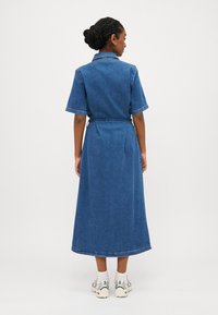 Denim obleka s kratkimi rokavi, ovratnikom in pasom. Obleka sega do sredine meč, z gladko teksturo in klasično modro barvo.