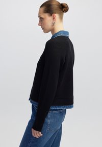 Zwarte geribbelde trui met lange mouwen, voorzien van een denim kraag die over een blauwe blouse is gelaagd. De algehele vorm is fitted en cropped.