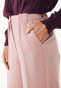 Persona che indossa pantaloni a vita alta di un pallido rosa con zip laterale e mano in tasca, abbinati a un maglione strutturato di un viola scuro.