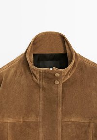 Bruine suède jas met hoge kraag, ritssluiting aan de voorkant die wordt bedekt door een klep met drukknoopjes, en een zwarte binnenvoering die zichtbaar is bij de kraag.