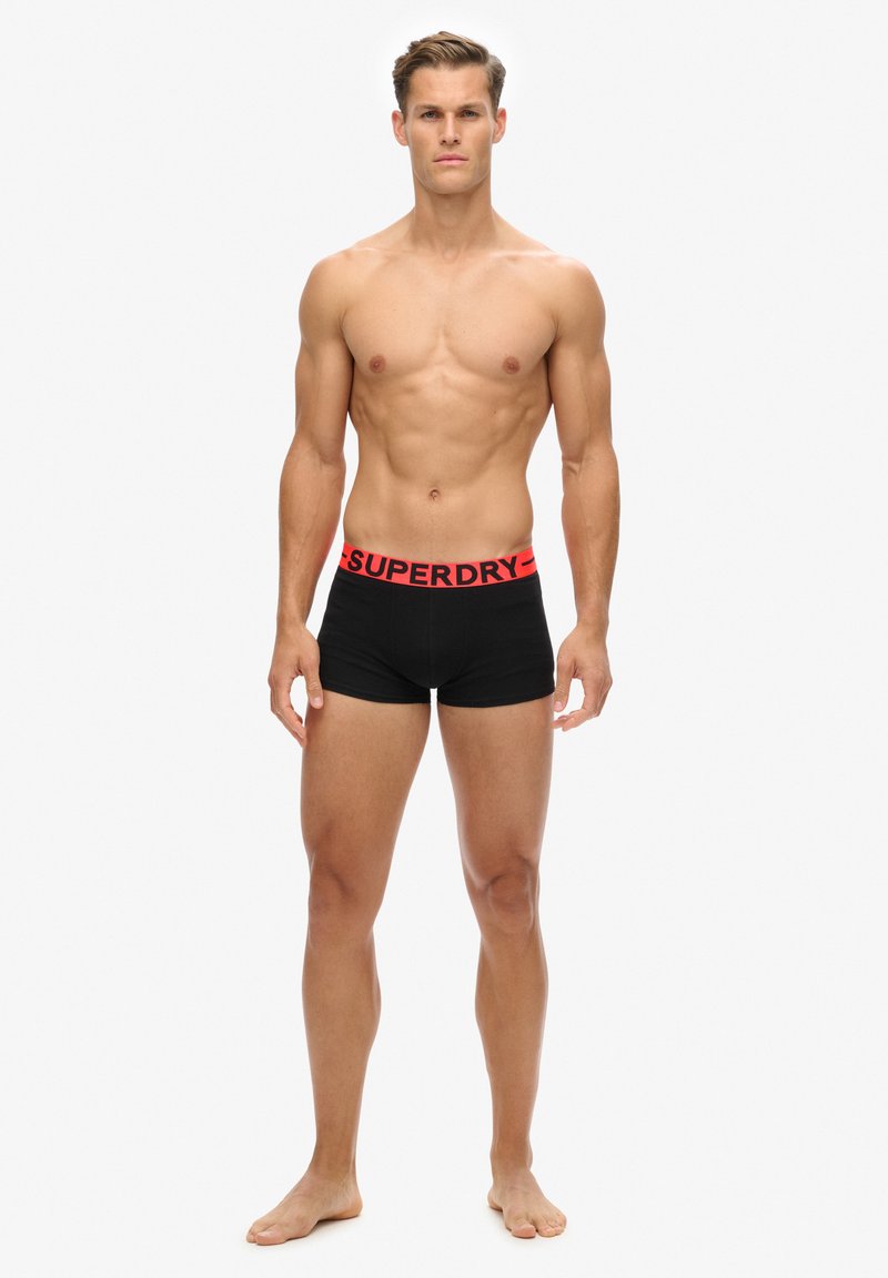 Boxer shorts noirs avec une ceinture rouge "SUPERDRY", affichant une coupe ajustée et une texture lisse, présentés sur un modèle masculin torse nu.