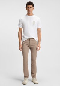 Tricou alb din bumbac cu o mică pată uzată pe piept, asociat cu pantaloni slim-fit din khaki și adidași crem.