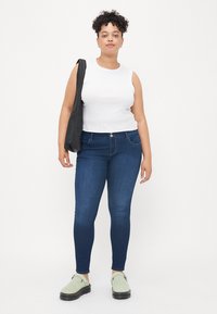 Modré džíny skinny, směs bavlny, střední výška pasu s předními kapsami, v kombinaci s bílým topem bez rukávů a světle zelenými botami. Černá kabelka.