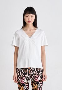 Witte katoenen t-shirt met korte mouwen en een v-hals, met een textuurpatroon van een vlinder op de voorkant. Gecombineerd met een patroonlegging.