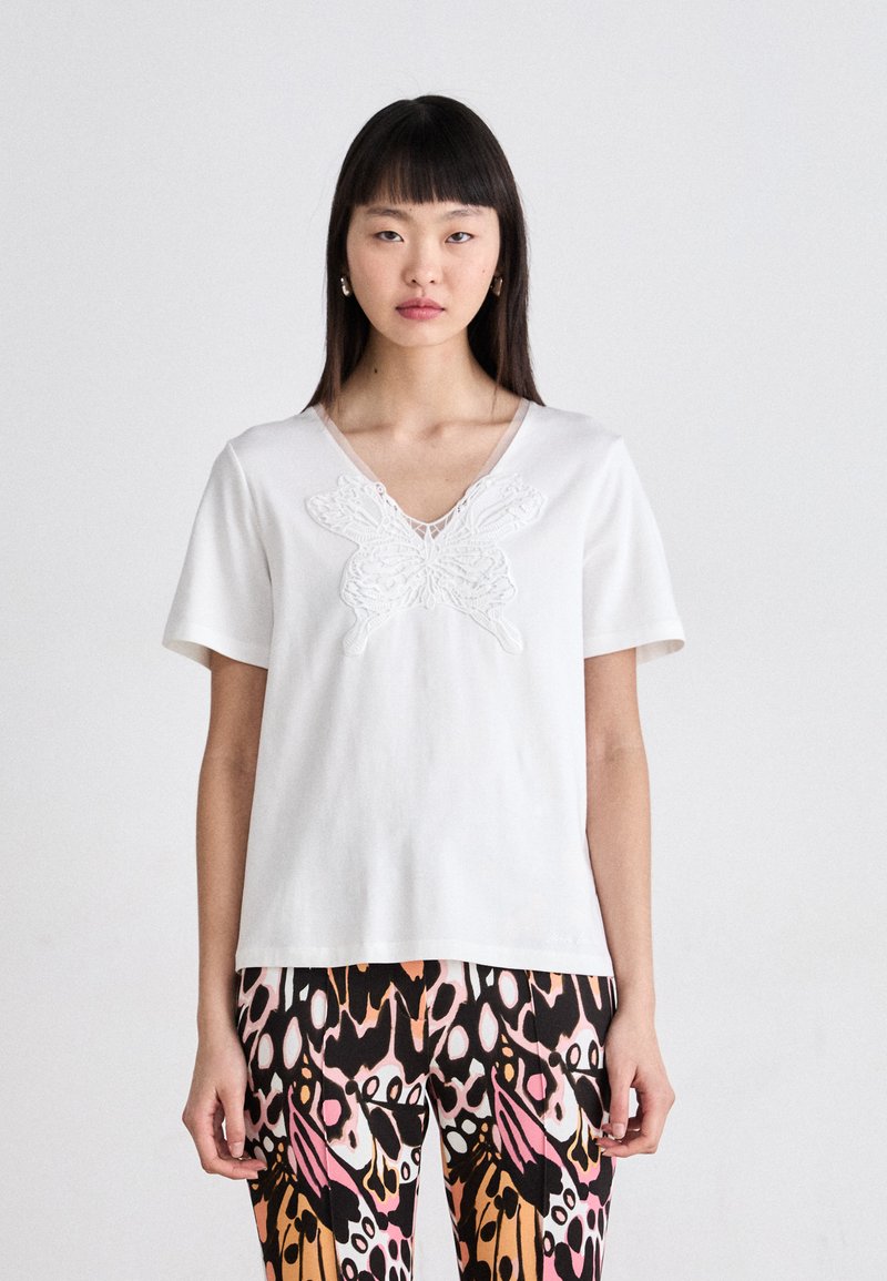 Witte katoenen t-shirt met korte mouwen en een v-hals, met een textuurpatroon van een vlinder op de voorkant. Gecombineerd met een patroonlegging.