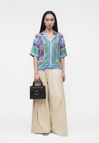 Femme portant une chemise à manches courtes à motifs bleu, vert et violet, pantalon large beige, tenant un sac à main noir avec un détail d'anneau doré.