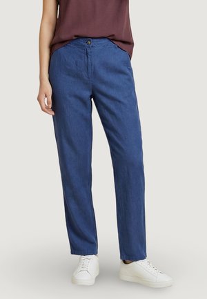 Persona che indossa pantaloni blu a gamba dritta, sneakers bianche e una maglia viola scuro infilata, in piedi contro uno sfondo chiaro.