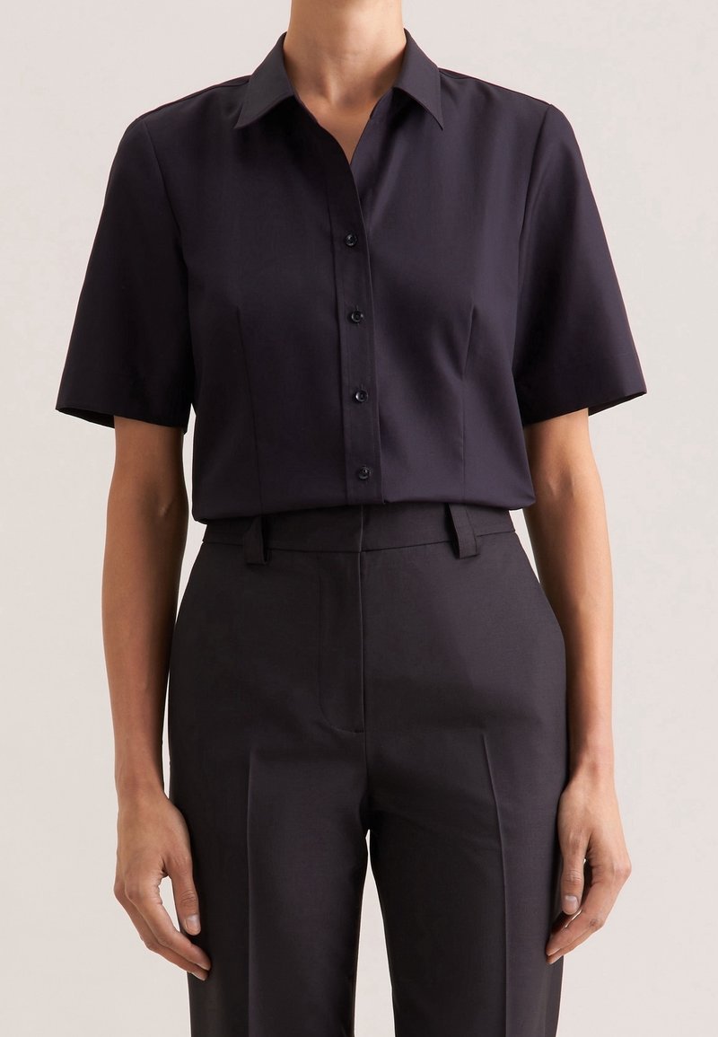Personne portant une chemise noire ajustée à manches courtes boutonnée, rentrée dans un pantalon noir taille haute, sur un fond uni.