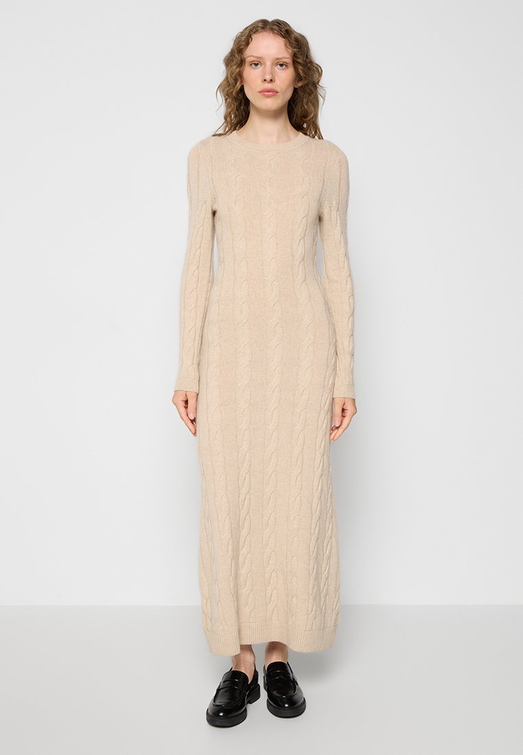 Beiges Kabelstrickkleid mit langen Ärmeln, tailliertem Schnitt, geripptem Saum und getragen mit schwarzen Slippern.