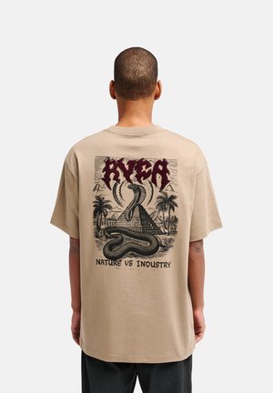 Persona che indossa una t-shirt beige con una grafica sul retro di un serpente, una piramide, palme e il testo "RVCA" e "Nature vs Industry."
