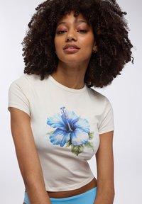 Crop top color crema con maniche corte, caratterizzato da un motivo di fiore hibiscus blu e foglie verdi. Tessuto dalla superficie liscia, vestibilità casual.