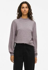 Maglione viola chiaro con collo alto, maniche lunghe a sbuffo e una consistenza morbida. Abbinato a pantaloni neri a vita alta.