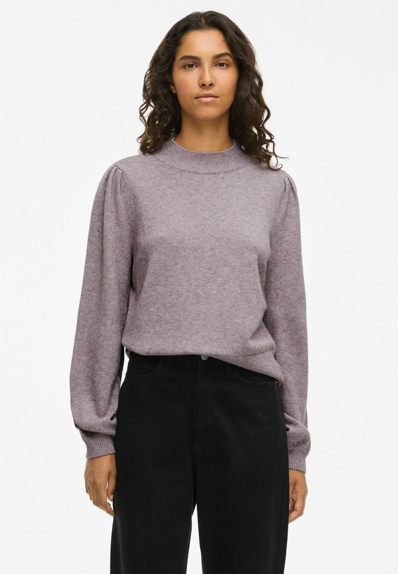 Maglione viola chiaro con collo alto, maniche lunghe a sbuffo e una consistenza morbida. Abbinato a pantaloni neri a vita alta.