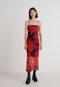 Vestido vermelho com estampado floral, com alças finas, ajustado ao corpete e uma saia fluida e transparente que cai até os tornozelos. Botas de plataforma pretas.