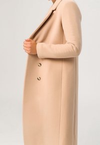 Femme portant un manteau de laine beige à double boutonnage, tenant le col d'une main, vue de l'épaule jusqu'à mi-mollet sur un fond uni.