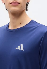 Νεαρό άτομο που φοράει μπλε αθλητική μπλούζα με λευκό τριγωνικό λογότυπο στο στήθος, εμφανίζεται από τον λαιμό μέχρι το πάνω μέρος του κορμού.