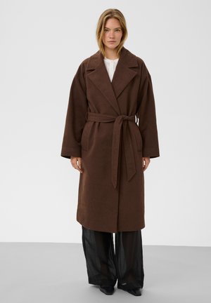Femme debout portant un long manteau marron ceinturé par-dessus un haut blanc et un pantalon large noir sur un fond clair uni.